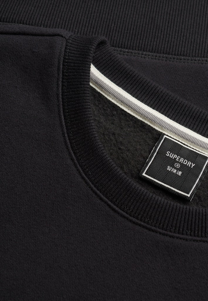 Close-up of a black crewneck sweatshirt showing the ribbed collar, a white stripe detail inside the neckline, and a black Superdry brand label.