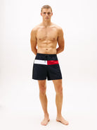 A shirtless male model stands against a plain white studio background wearing navy swim shorts with a white and red horizontal stripe detail at the waist.