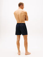 A man shown from the back wearing dark navy blue swim shorts, standing against a plain white studio background.