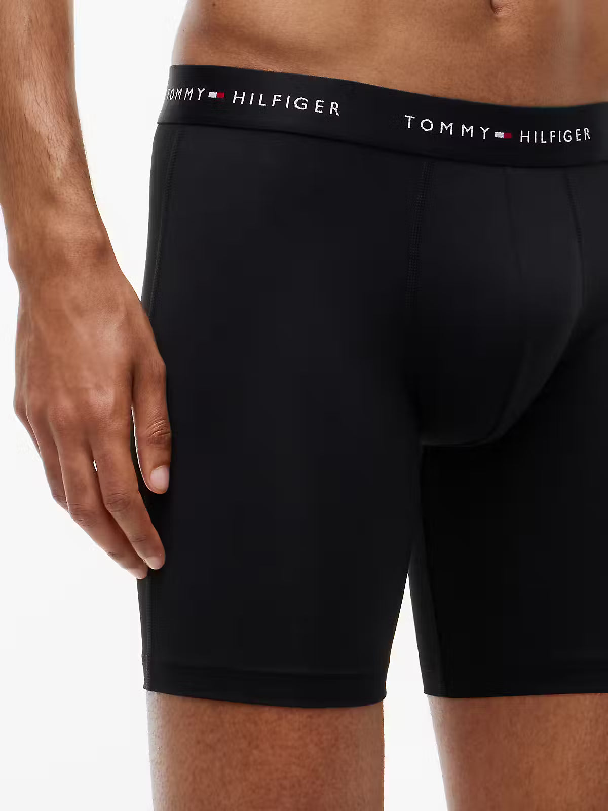 A close-up of a model wearing black Tommy Hilfiger boxer briefs, showing the branded elastic waistband and the fit of the underwear.